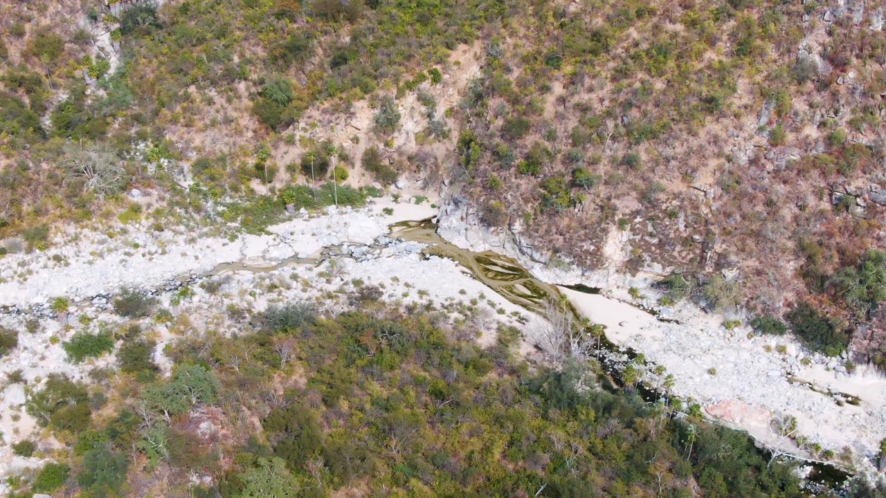 Overhead San Dionisio frame showing a diagonal rocky wash, a dark stream edge, dense green scrub, and dry brown slope vegetation, no visible people.