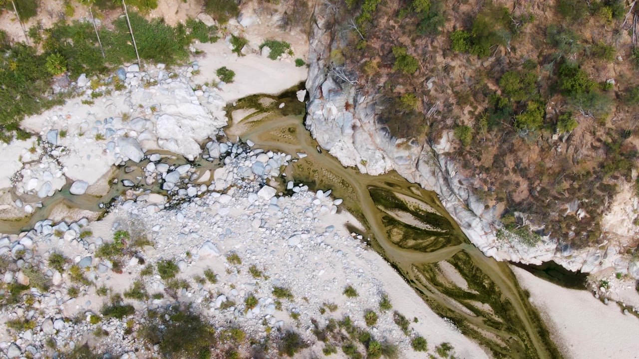 Aerial frame from San Dionisio showing shallow green-brown water curving past white boulders, a dry rocky hillside, sparse plants, and no visible people.
