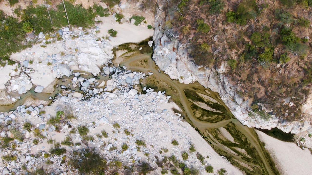 Top-down San Dionisio view with a narrow meandering water channel, white rock deposits, tan dry ground, scrub vegetation, and no visible people.