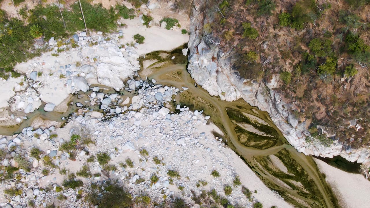 Overhead view in San Dionisio of a river bend bordered by white stones, dry hillside vegetation, exposed rock, sandy patches, and no visible people.
