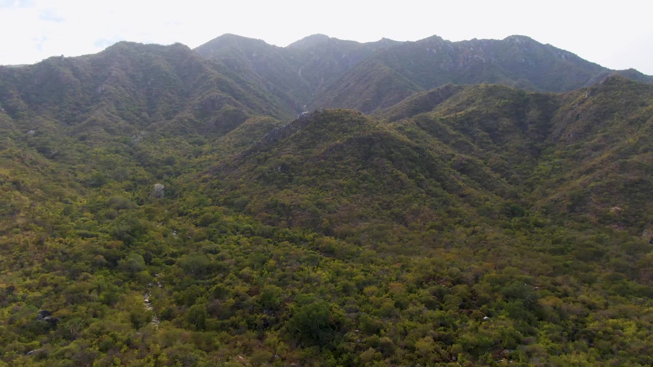 Wide San Dionisio aerial view with layered green and brown hills, a vegetated valley floor, small pale stream lines, bright overcast sky, and no visible people.