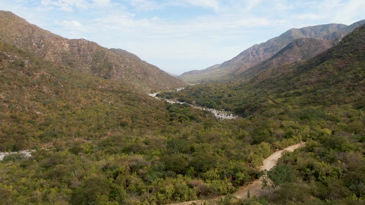 Early frame over San Dionisio valley terrain, with the curving dirt road more apparent in the lower foreground, scrubby vegetation across the valley floor, rocky mountains on both sides, and no visible people.
