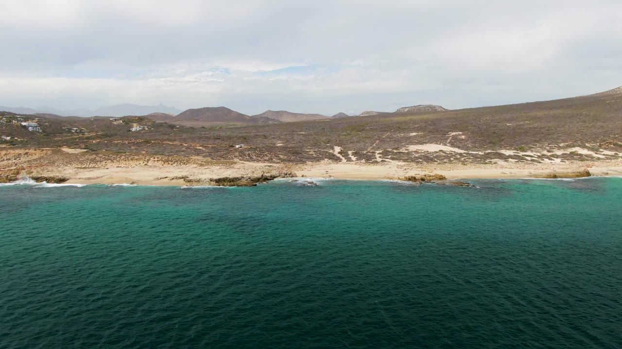 Aerial view at Shipwrecks, Baja California Sur, with deep teal water in front, a sandy beach and rocks along shore, low hills beyond, and no visible people.