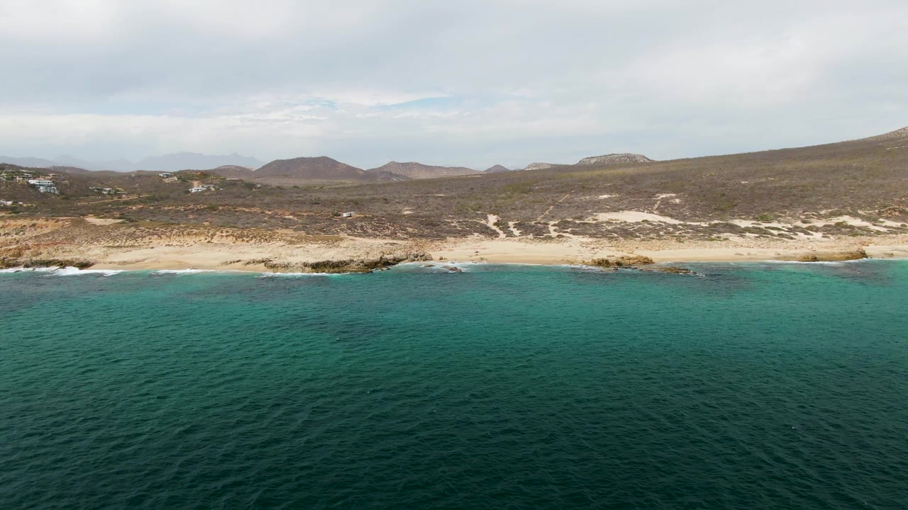 Turquoise surf reaches a sandy beach with rocky points and low brown hills behind it.