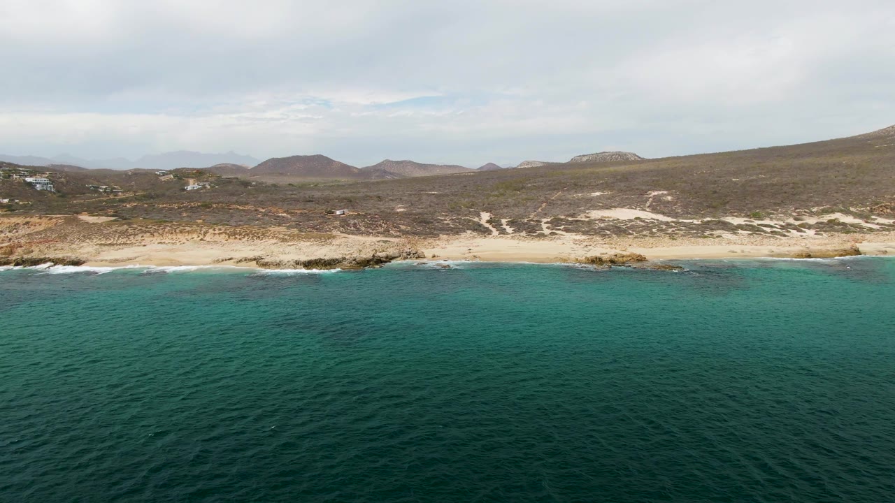 Wide aerial coastline at Shipwrecks shows dark ocean in the foreground, pale sand broken by rocks, hazy hills in the background, and no visible people on the shore.