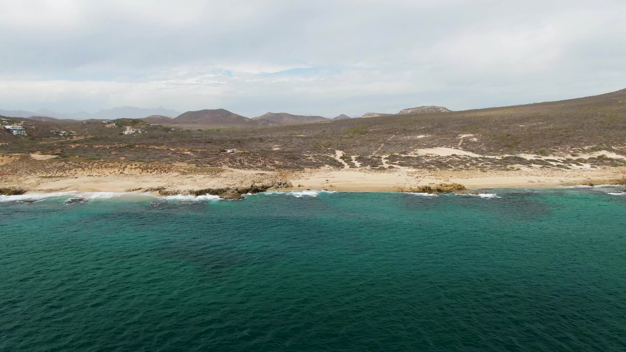 Aerial frame over Shipwrecks East Cape in Baja California Sur showing turquoise ocean in front of a sandy beach, rocky shoreline, low scrub hills, cloudy sky, and no visible people.