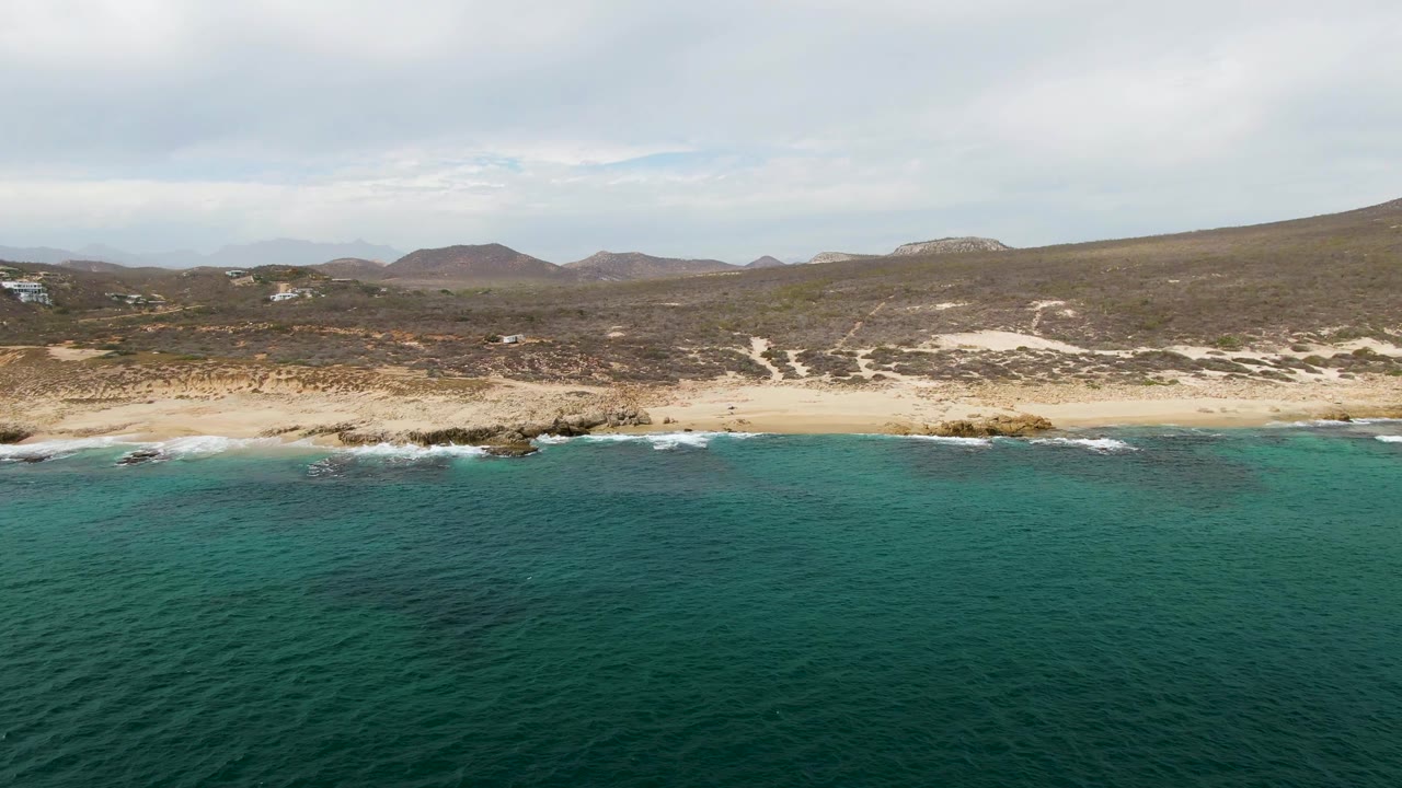Aerial view at Shipwrecks East Cape with darker turquoise water in the foreground, a pale sand beach across the center, rocky breaks in the surf, low hills behind, and no visible people.