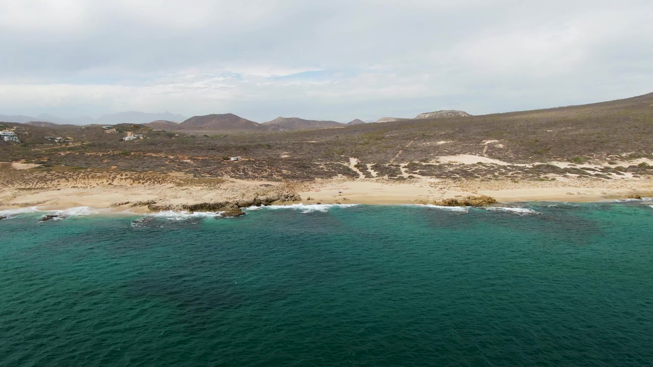Aerial frame from Shipwrecks East Cape showing gentle waves reaching a sandy shoreline with several rock clusters, dry hills beyond the beach, soft cloud light, and no visible people.