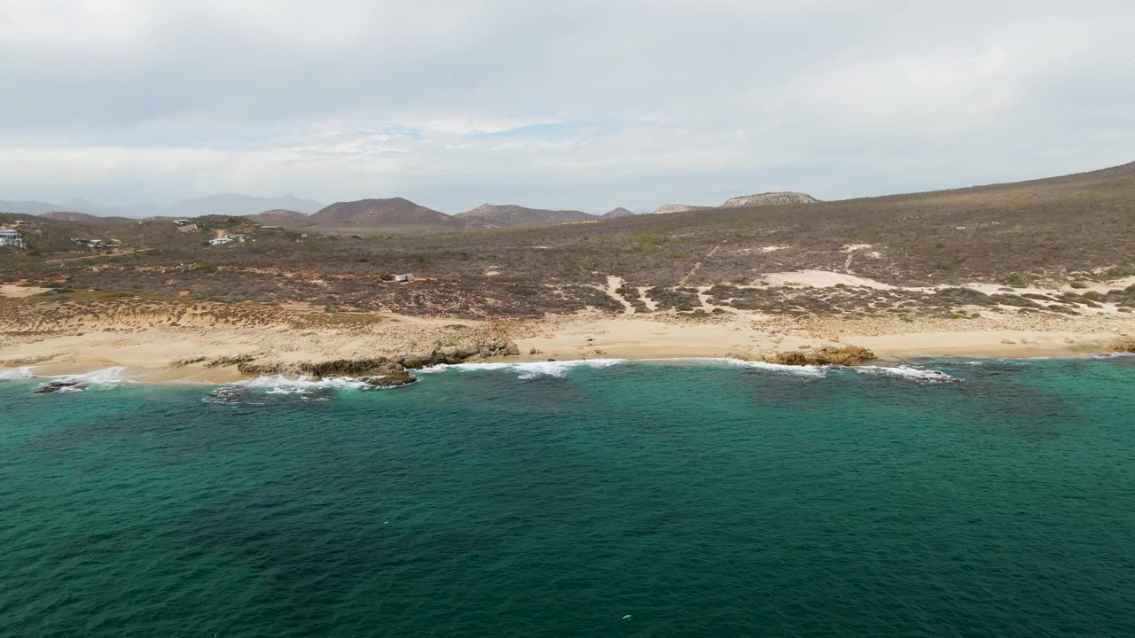 Mid-clip aerial view at Shipwrecks East Cape with textured green-blue ocean in front, a thin surf line along the sand, rocky points on the beach, distant hills, and no visible people.