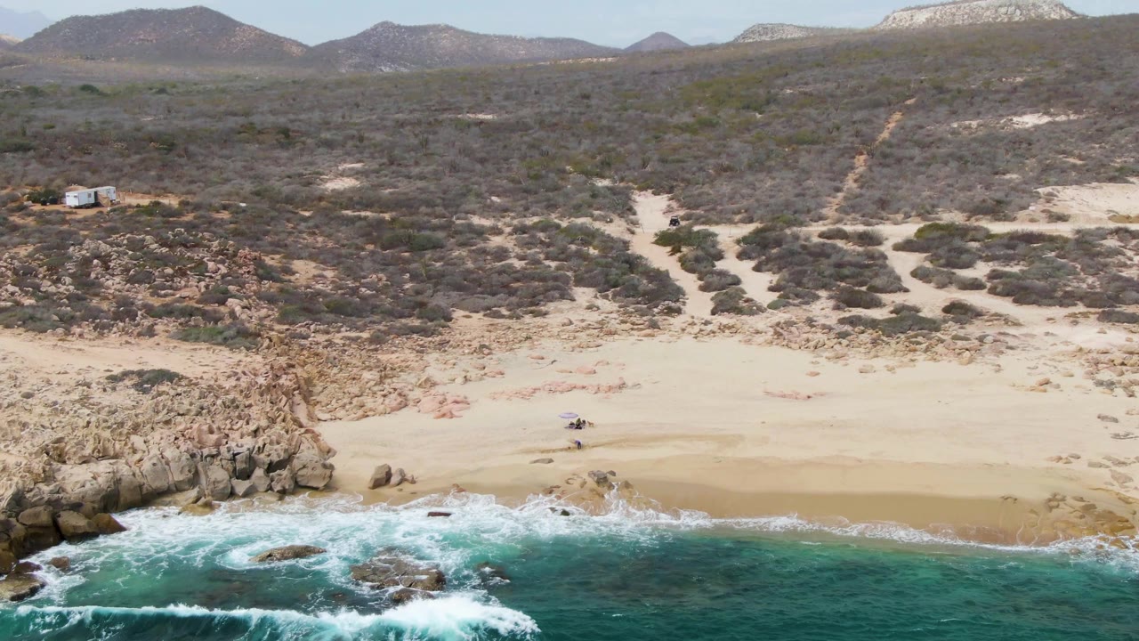 Aerial frame over Shipwrecks, Baja California Sur showing teal waves at the bottom edge, a broad sandy beach, a central rocky outcrop, scrub hills, distant small structures, and tiny people near the shoreline.