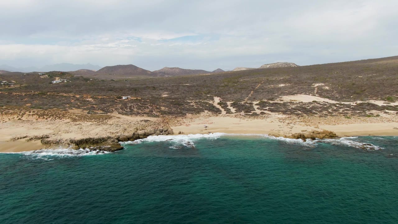 Aerial frame over Shipwrecks, Baja California Sur, with turquoise ocean in front, pale sand and rocky outcrops along the shore, scrub hills behind, and no visible people at this moment.