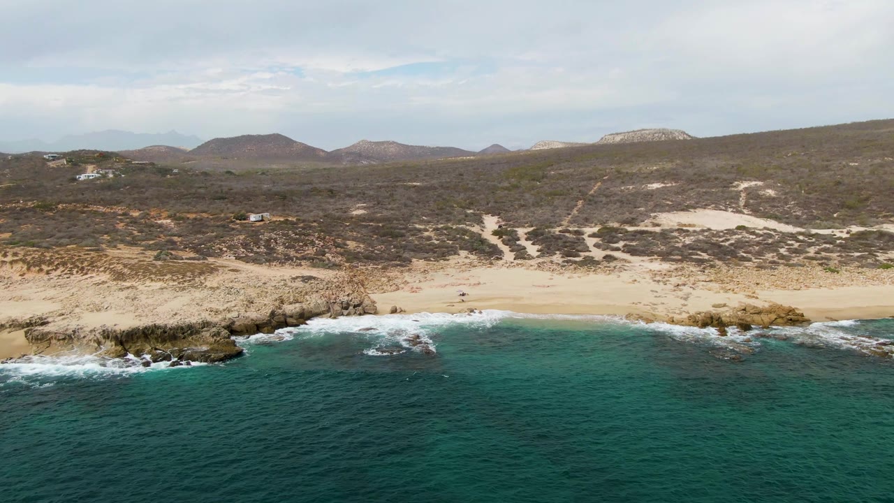 Wide aerial shoreline view with waves breaking on rocks and low hills behind the beach.