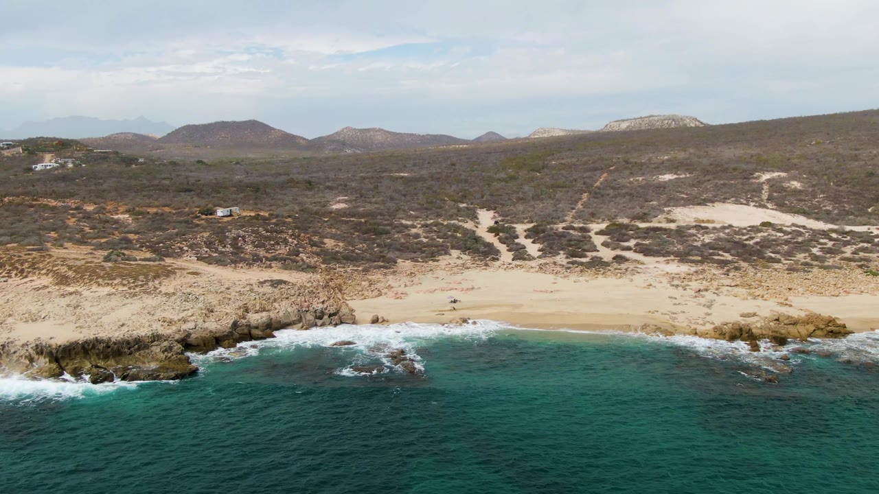 Shipwrecks, Baja California Sur shoreline frame with white surf curling around rocky outcrops, pale sand behind it, scattered tiny people on the beach, and scrubby hills under light cloud.