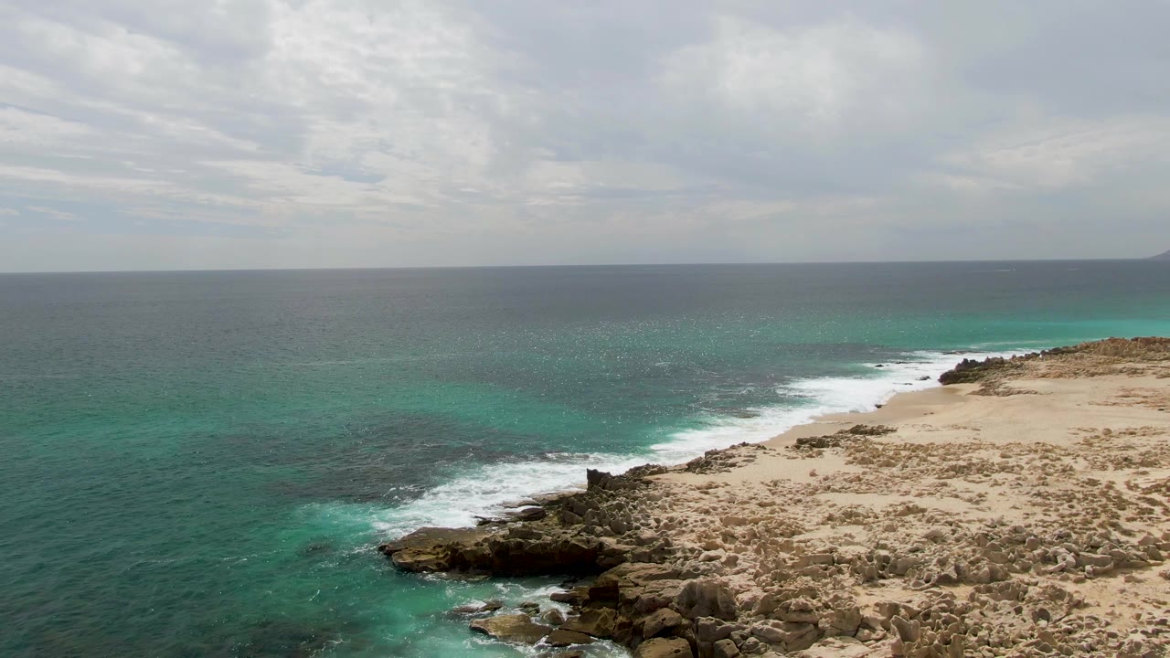 Aerial view of waves breaking along a rocky coastline beside turquoise water.