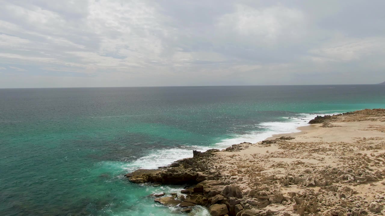 Aerial view at Shipwrecks, Baja California Sur with whitewater curling around the rocky point, deeper blue-green water offshore, dry shoreline terrain, and no visible people.