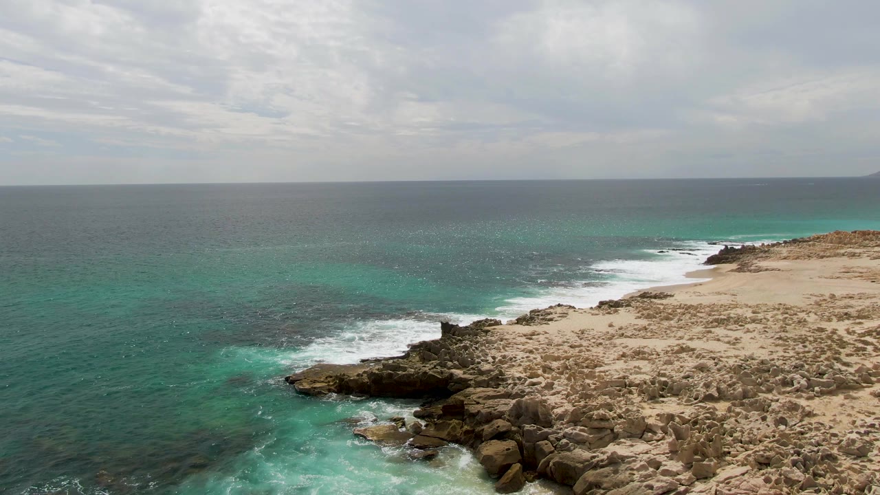 Aerial frame over Shipwrecks, Baja California Sur showing a broader section of sandy, rocky coast, turquoise shallows, white surf bands, cloud cover, and no visible people.