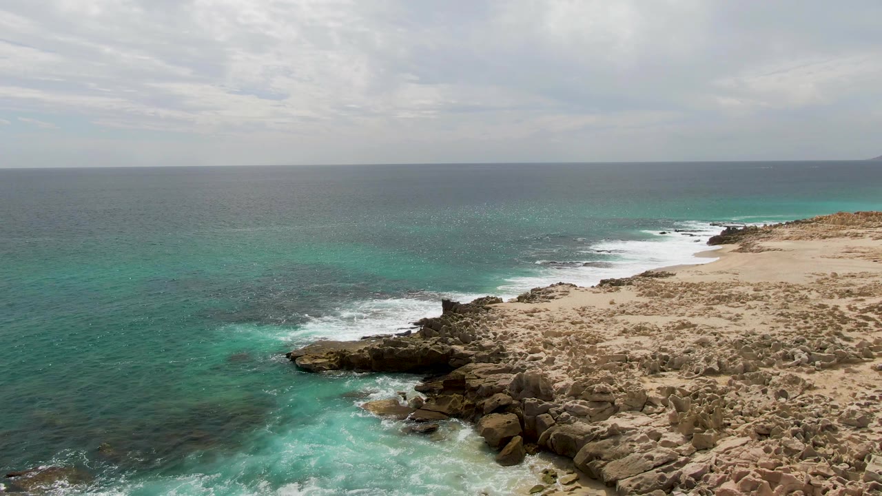 Aerial view at Shipwrecks, Baja California Sur with the rocky outcrop lower in frame, pale sand near the surf, blue-green ocean extending left, and no visible people.