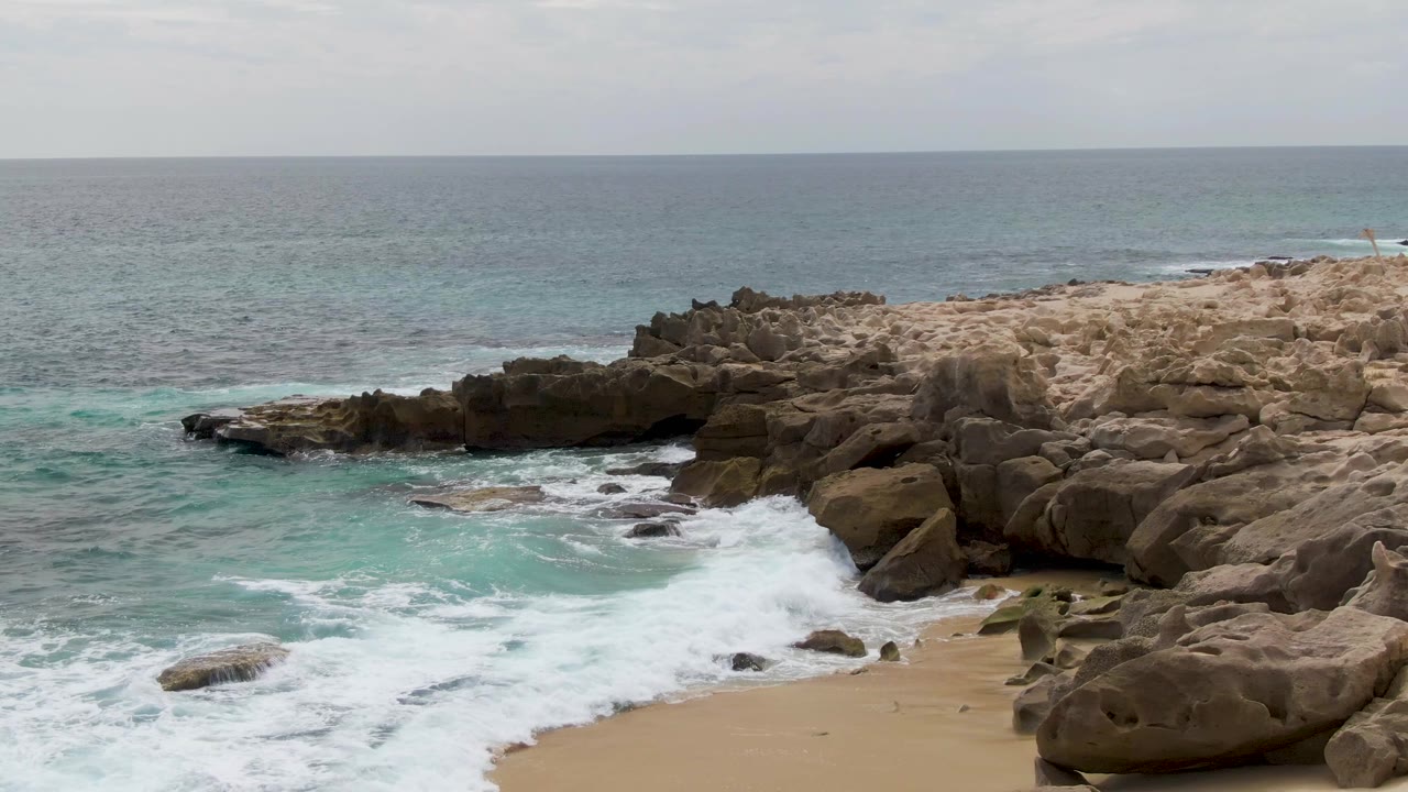 At Shipwrecks in Baja California Sur, turquoise surf breaks against tan shoreline rocks with a small strip of sand at lower right and no visible people.