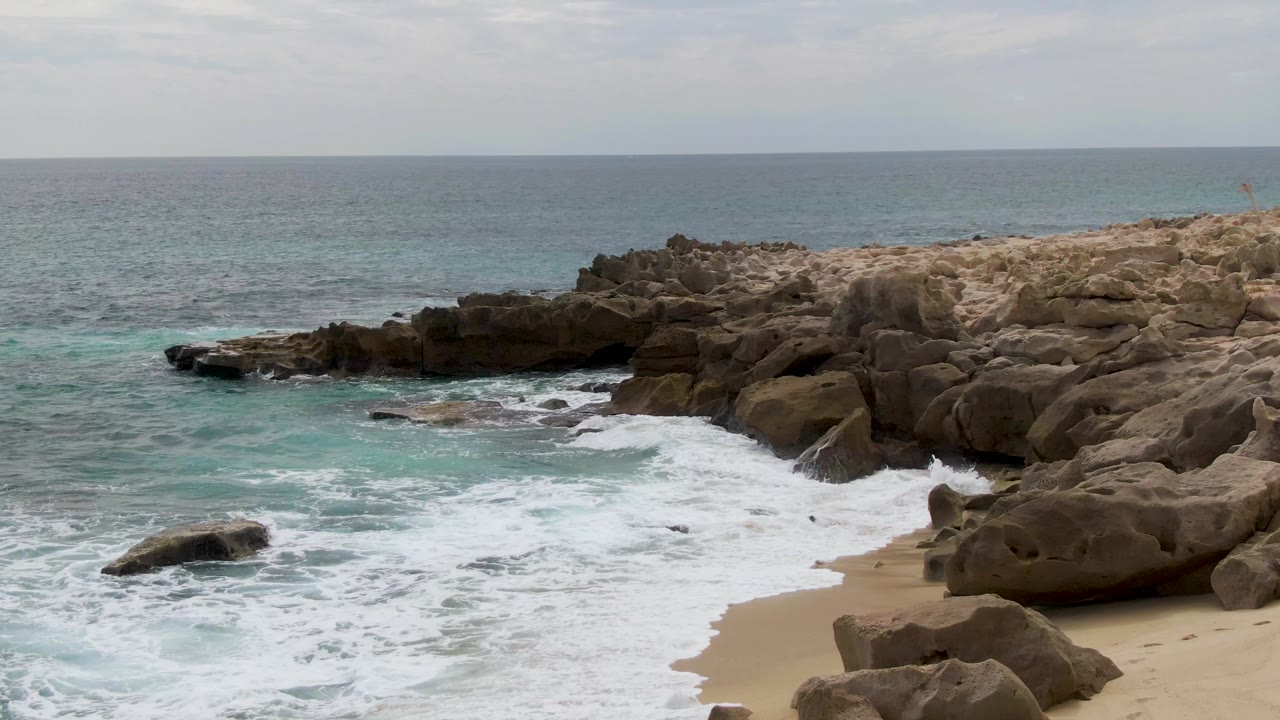 At Shipwrecks in Baja California Sur, foamy water spreads across the sand beside rounded eroded rocks, with open ocean beyond and no visible people.