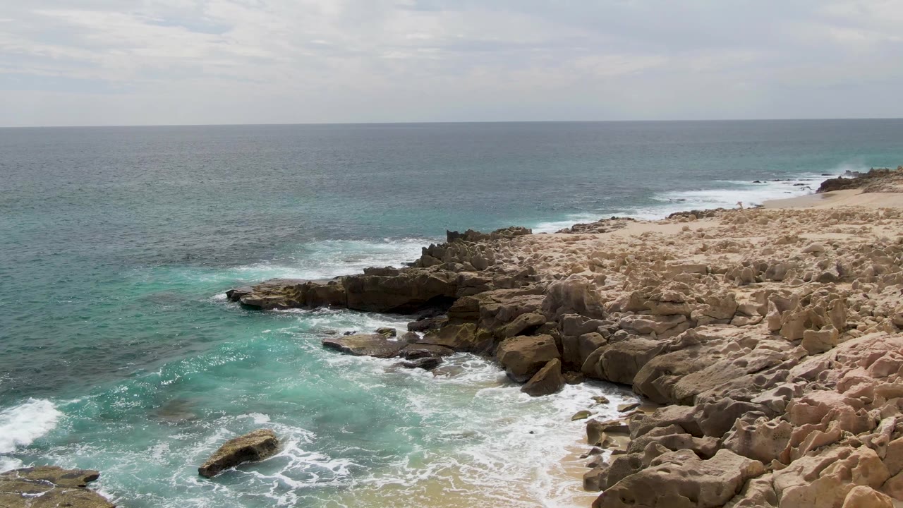 Aerial frame at Shipwrecks, East Cape, Baja California Sur, with white foam curling around a dark offshore rock and tan shoreline rocks, with no visible people.
