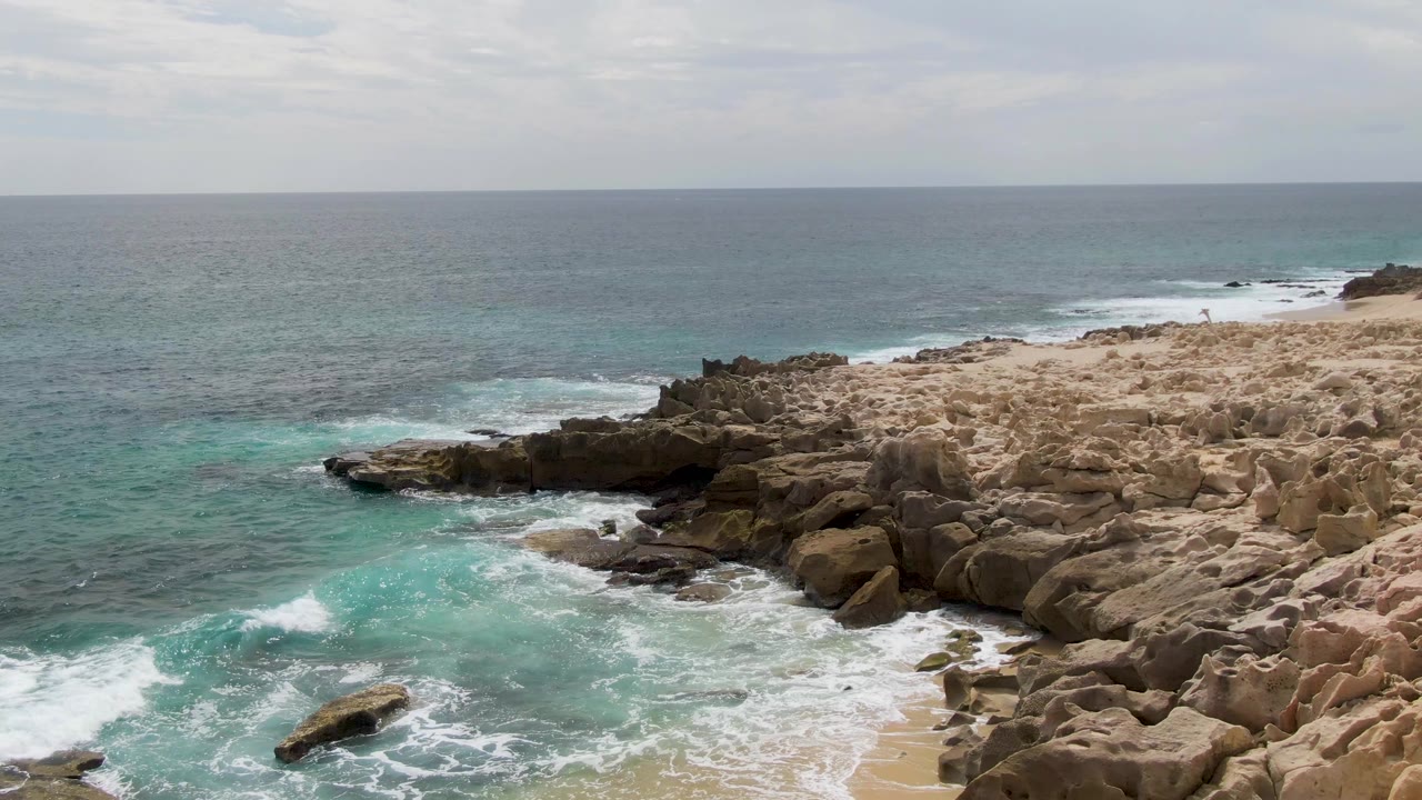 Aerial view at Shipwrecks, East Cape, Baja California Sur, where a brighter turquoise patch spreads beside jagged tan rocks under flat overcast light, with no visible people.