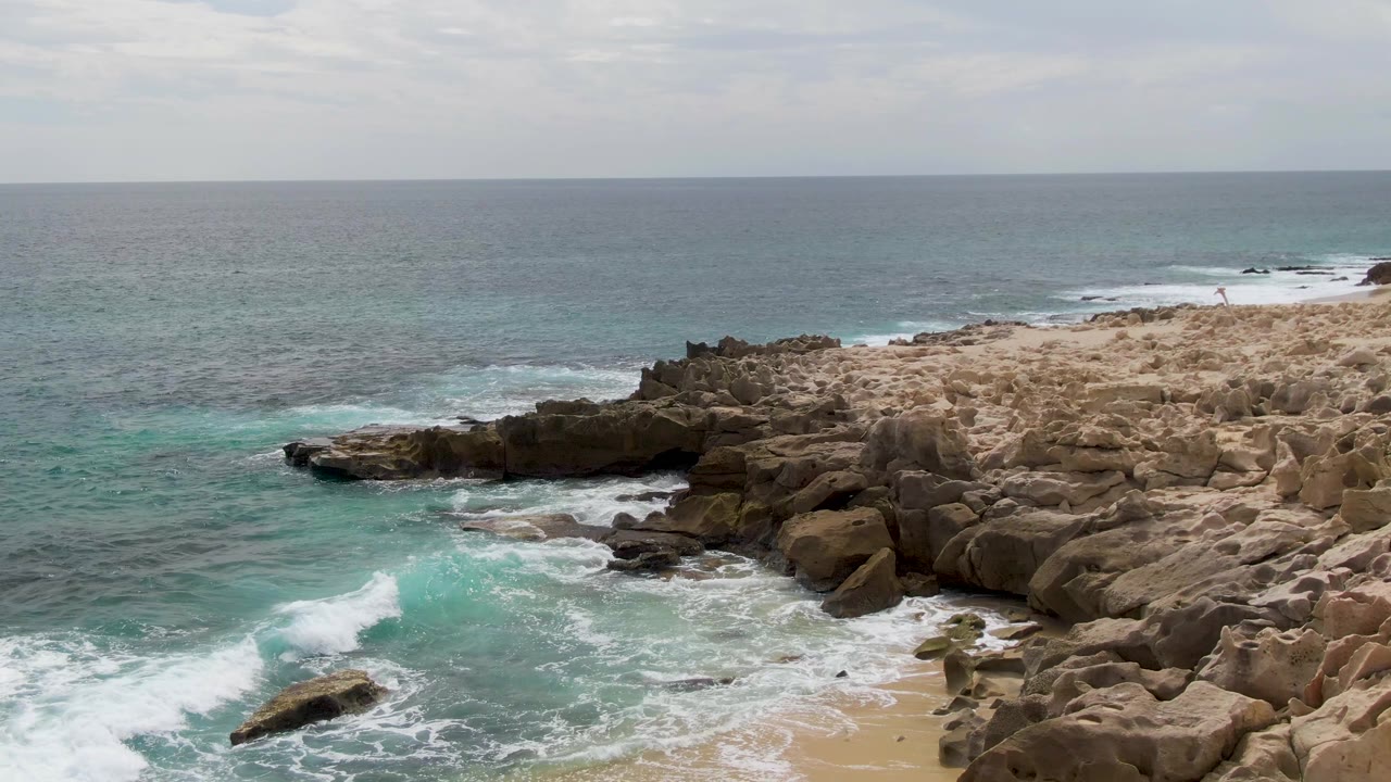 Aerial frame at Shipwrecks, East Cape, Baja California Sur, with surf spreading toward a small sandy strip below the rock formations and no visible people.