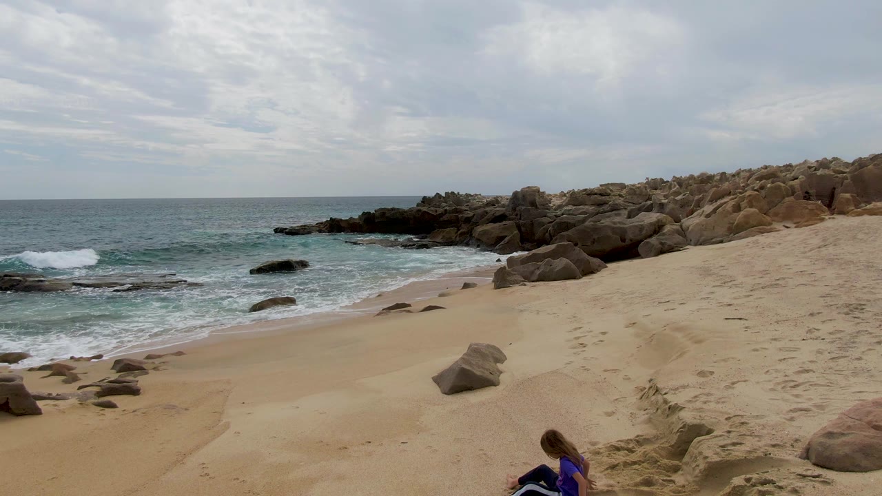 At Shipwrecks, Baja California Sur, a child in a blue shirt sits near the lower edge of a sandy beach while small waves reach the rocky shoreline under a cloudy sky.