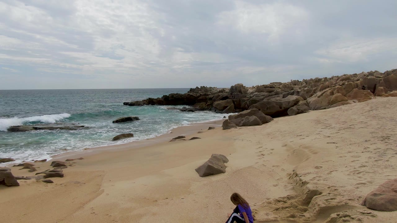 Wide beach view with a child near the lower edge, surf, rocks, and cloudy sky.