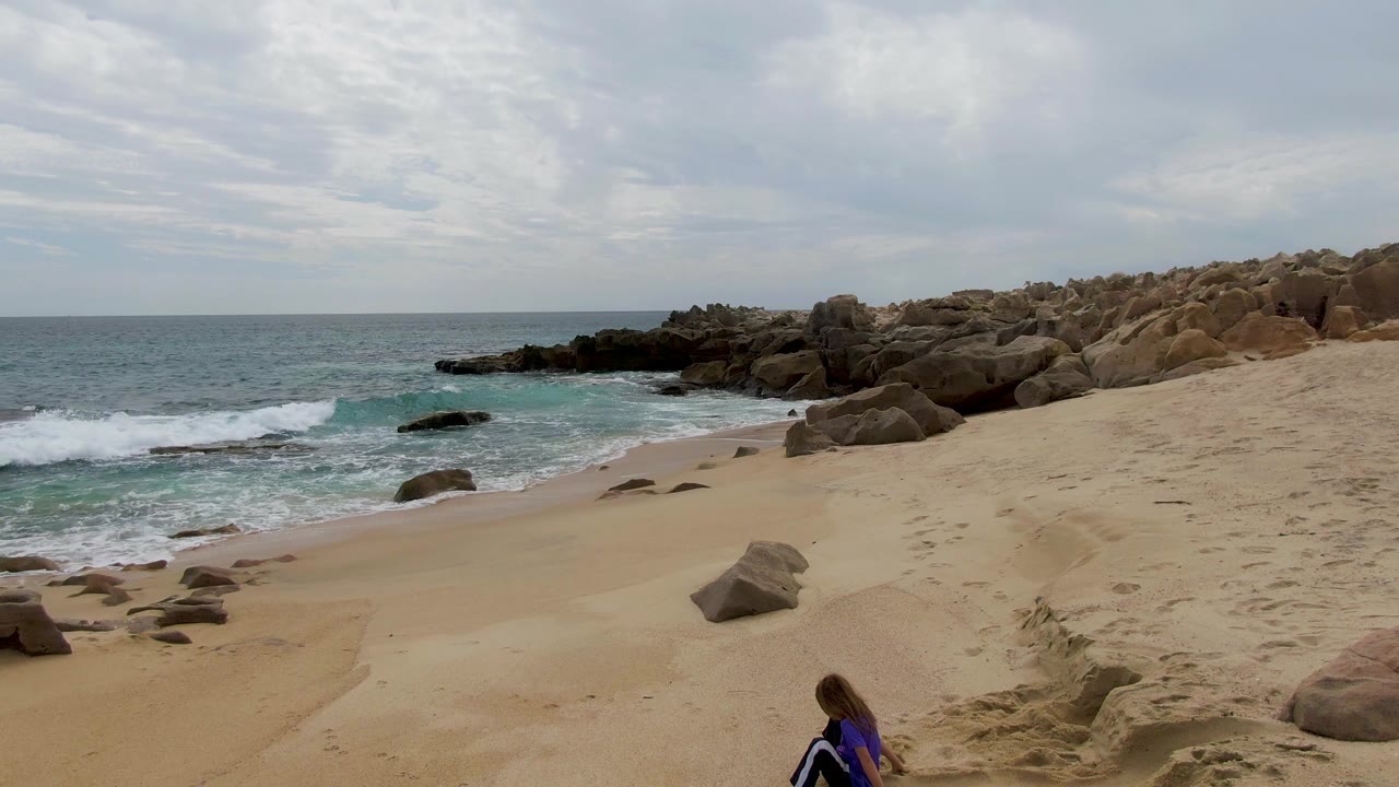 At Shipwrecks, Baja California Sur, the child is partly visible near the lower right while a white wave breaks against the left rocks and the sandy beach curves toward the ocean.