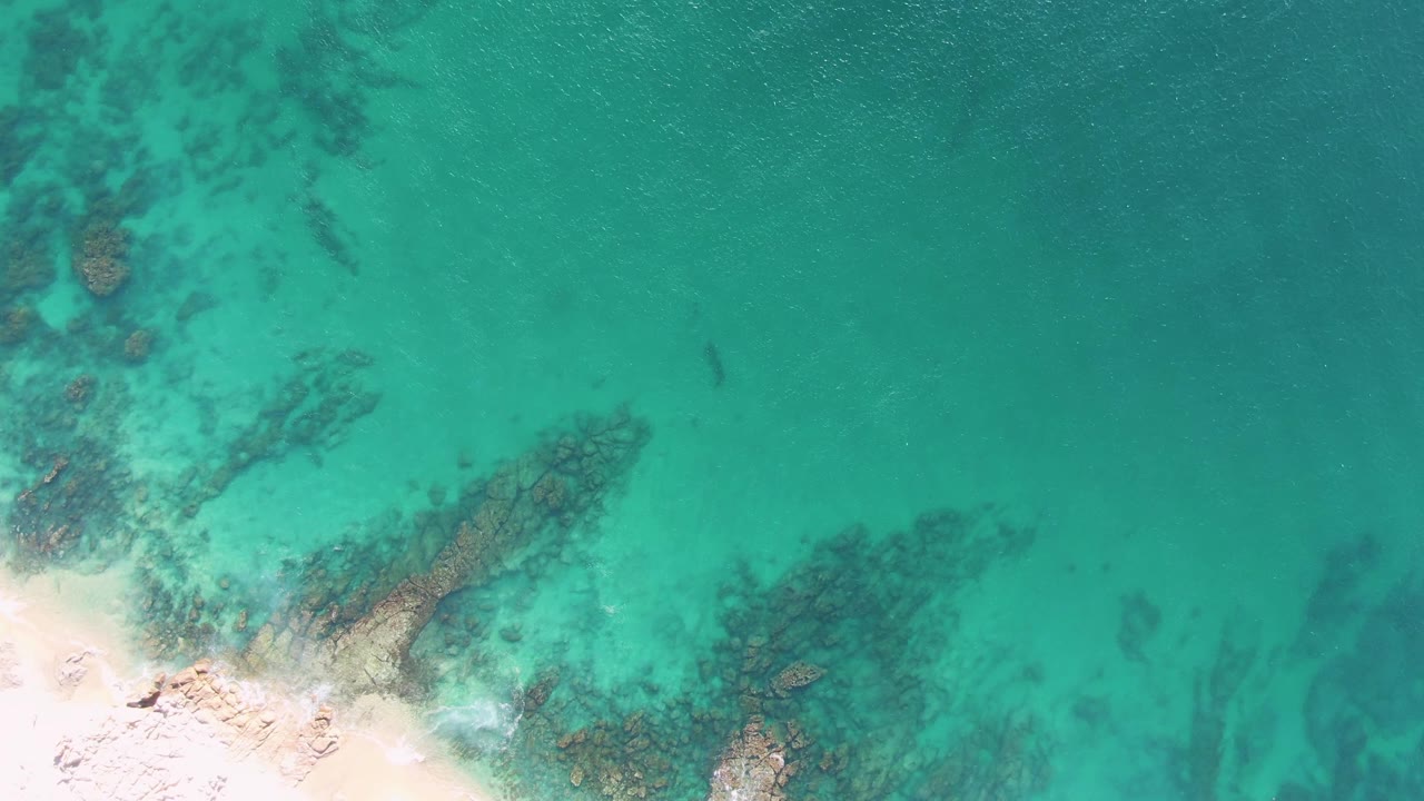 Overhead view of Buzzards Beach turquoise shallows with pale sand at lower left, dark submerged rocks through the water, faint wave wash, and no visible people.