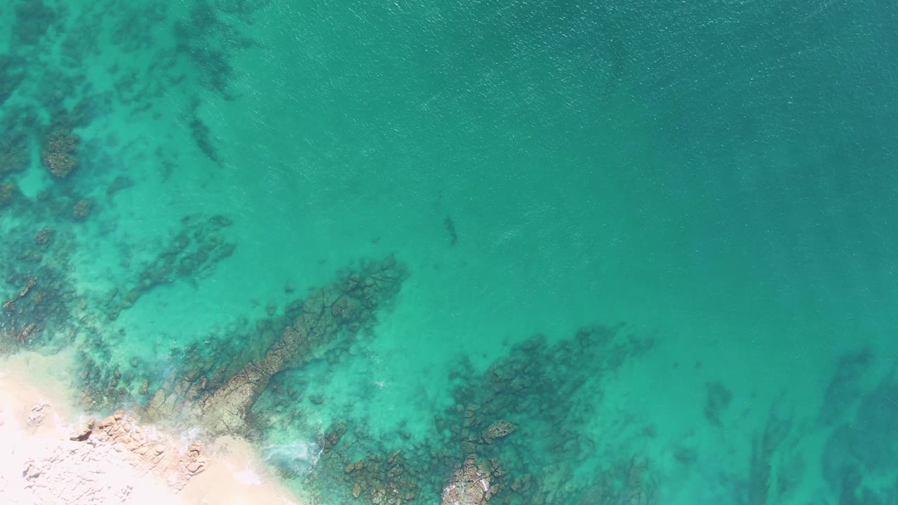 Buzzards Beach shallows from above, with a brighter sandy lower-left corner, dark rock clusters visible beneath green-blue water, and no visible people.