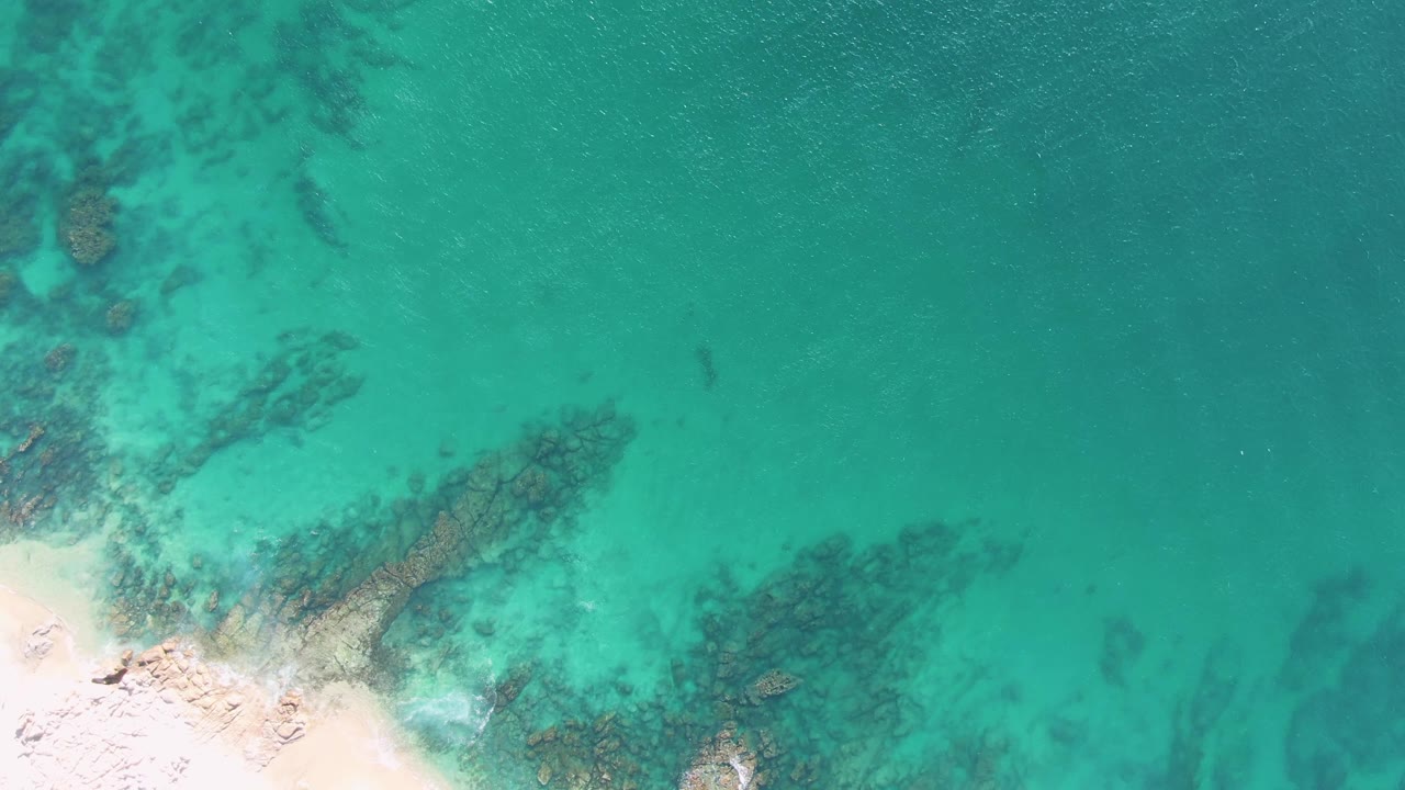 Overhead Buzzards Beach frame with the sandy shore partly cropped at lower left, underwater rock bands crossing the shallows, textured turquoise surface, and no visible people.