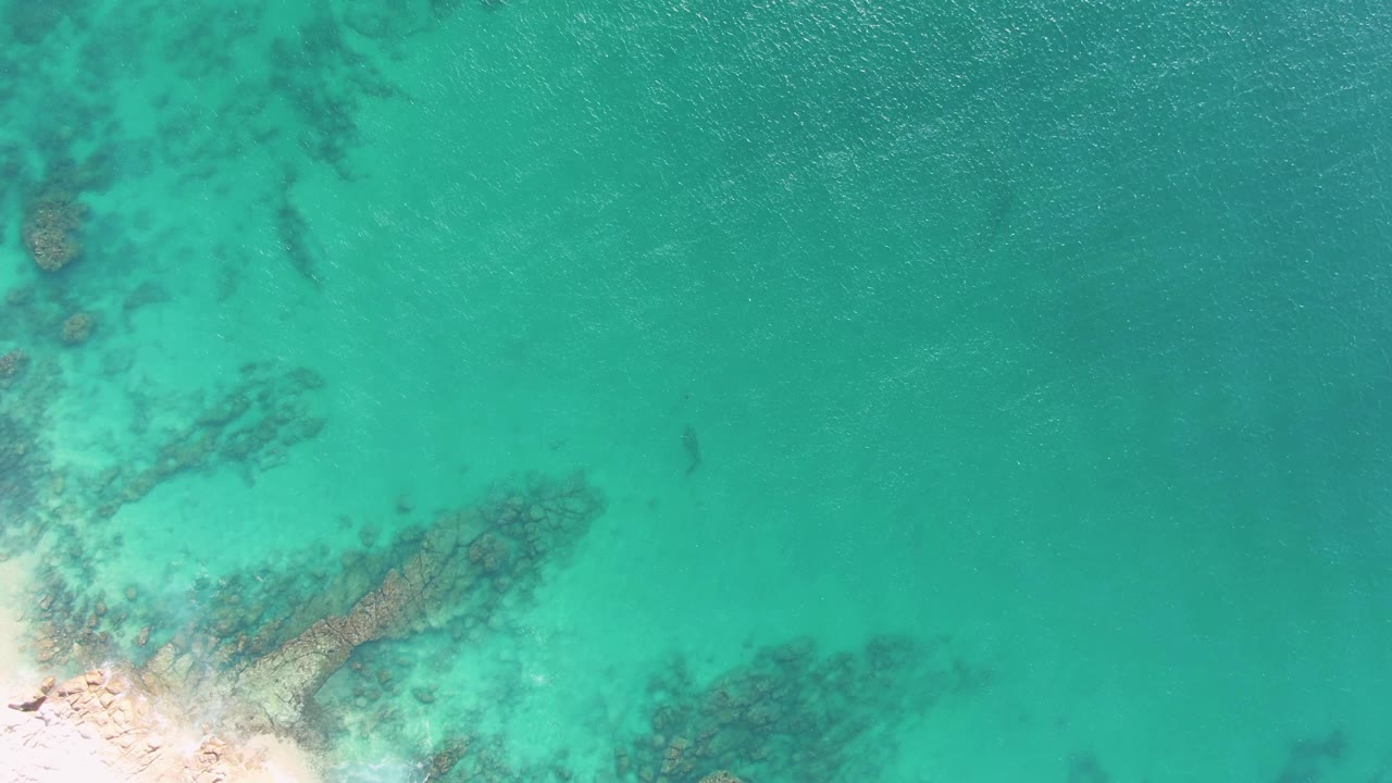 Overhead view of clear turquoise water at Buzzards Beach, Baja California Sur, with dark underwater rock patches near a sandy lower-left shoreline and no visible people.