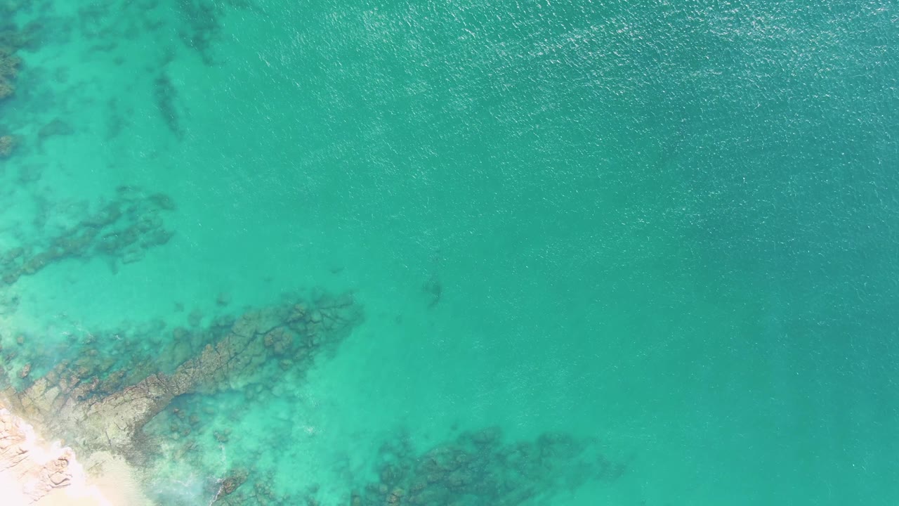 Overhead view at Buzzards Beach, Baja California Sur, showing turquoise shallows, dark submerged rocks, pale sand at the lower left, and no visible people.