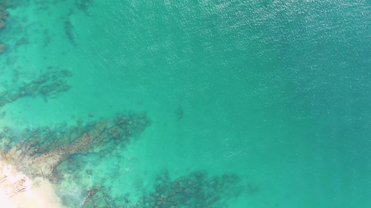 Second overhead frame from Buzzards Beach shows the sandy lower-left shore, a diagonal band of submerged rocks, clear turquoise water, and no visible people.