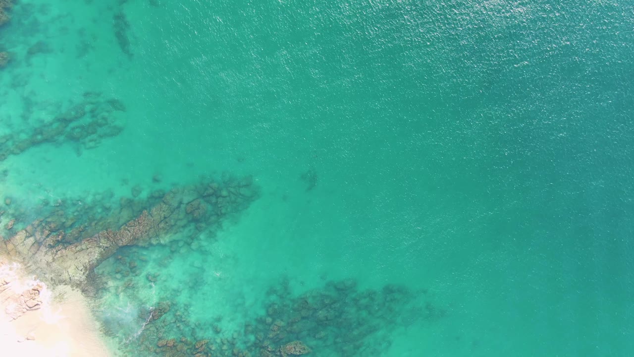 Overhead Buzzards Beach frame with more open turquoise water to the right, submerged rocks near the lower and left areas, pale sand at the lower left, and no visible people.