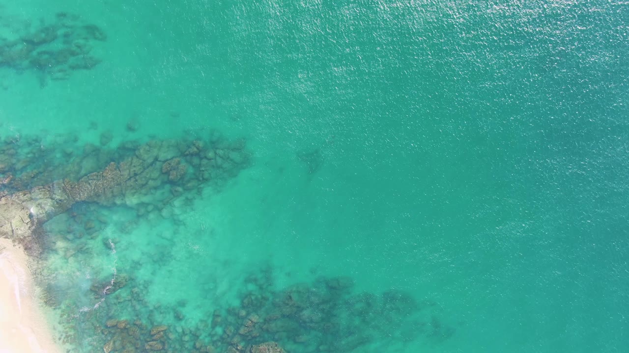 Top-down view at Buzzards Beach in Baja California Sur, Mexico, showing clear turquoise water, submerged rocks, a pale sandy shore along the left edge, and no visible people.