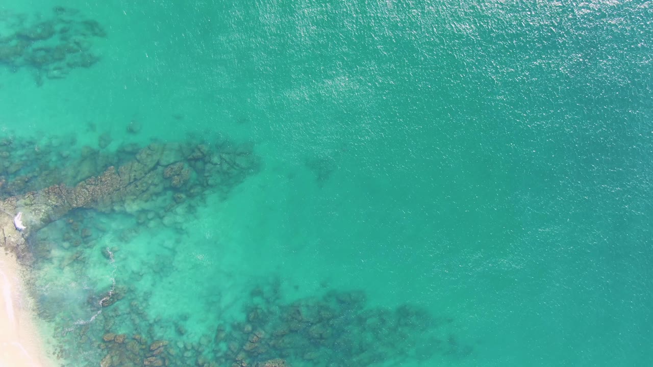 Aerial frame at Buzzards Beach in Baja California Sur, Mexico, with shallow turquoise water, underwater rock formations, a sliver of sand at left, and no visible people.