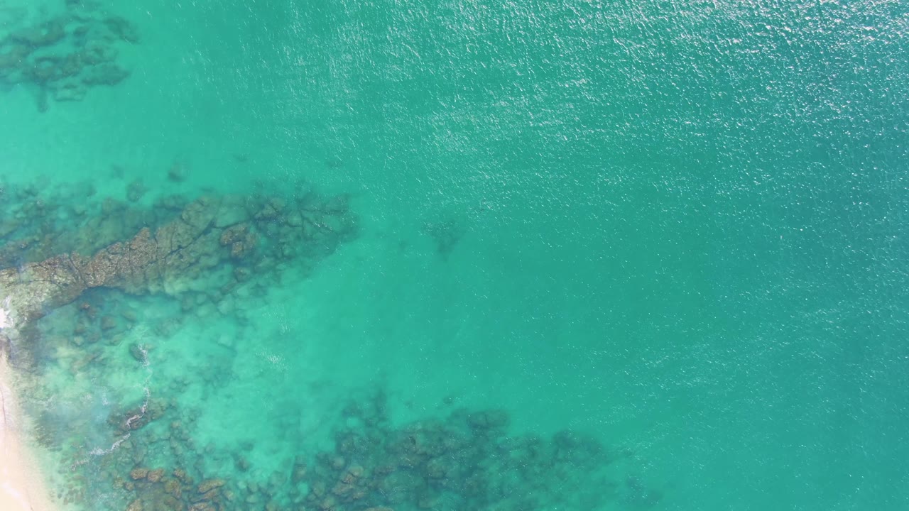 Overhead view at Buzzards Beach in Baja California Sur, Mexico, showing sunlit turquoise water, submerged rocky reef along the left and lower areas, and no visible people.