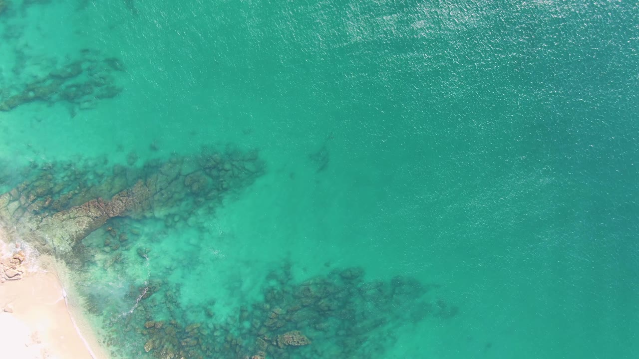 Overhead frame from Buzzards Beach in Baja California Sur showing pale sand at left, clear turquoise shallows, dark submerged rocks, surface ripples, and a small person on the shore.