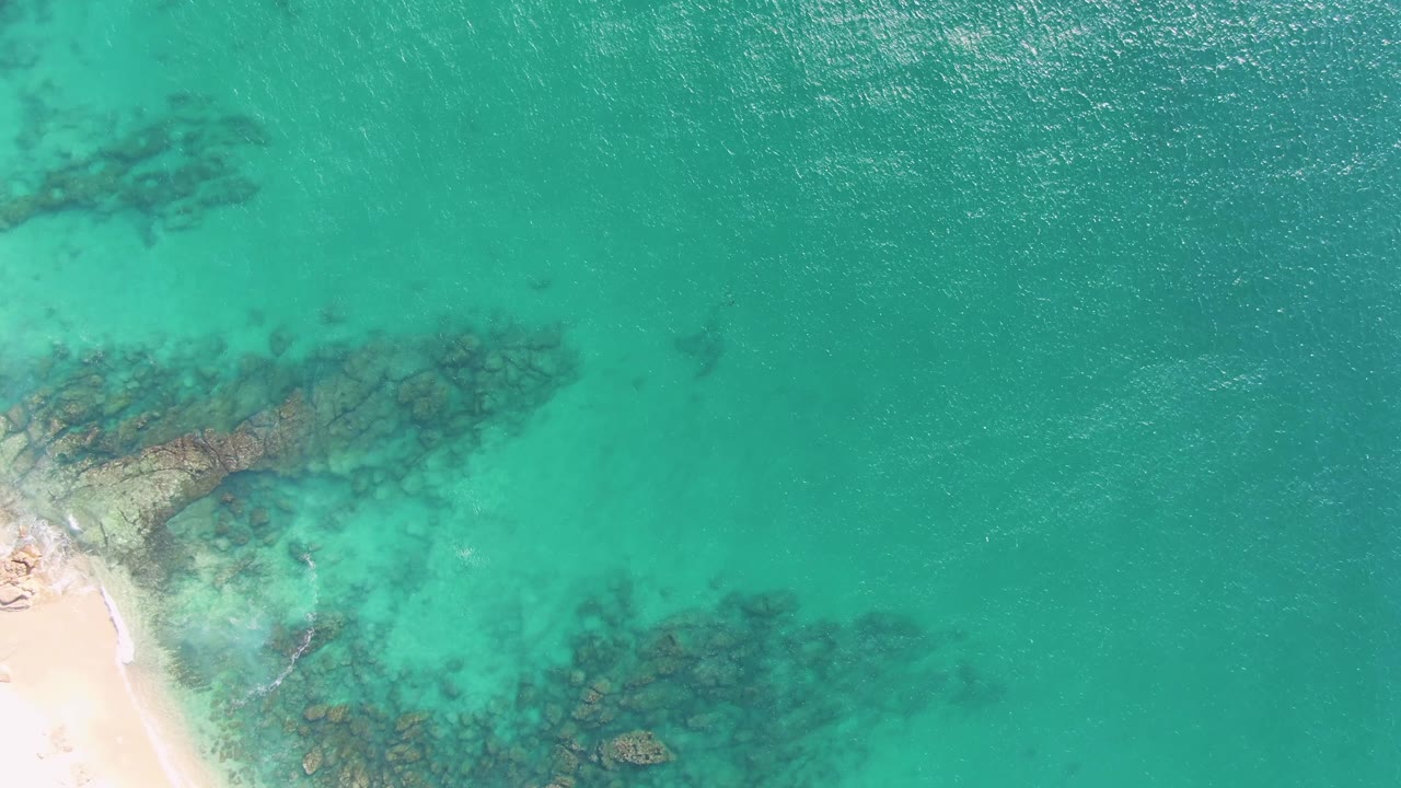Aerial view of turquoise ocean, sandy edge, and submerged rocks with no visible people.