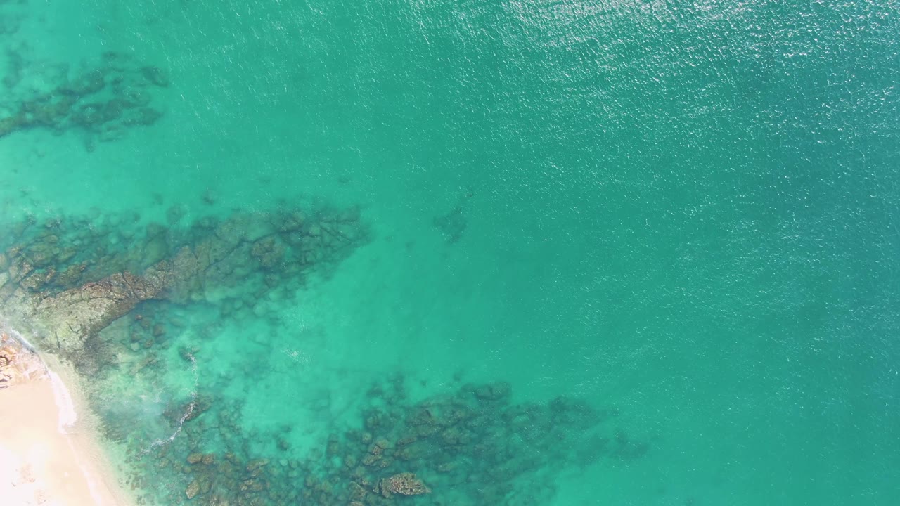 Buzzards Beach aerial frame showing rippled turquoise sea across most of the view, a sliver of pale sand at left, and clustered submerged rocks in the shallows with no visible people.