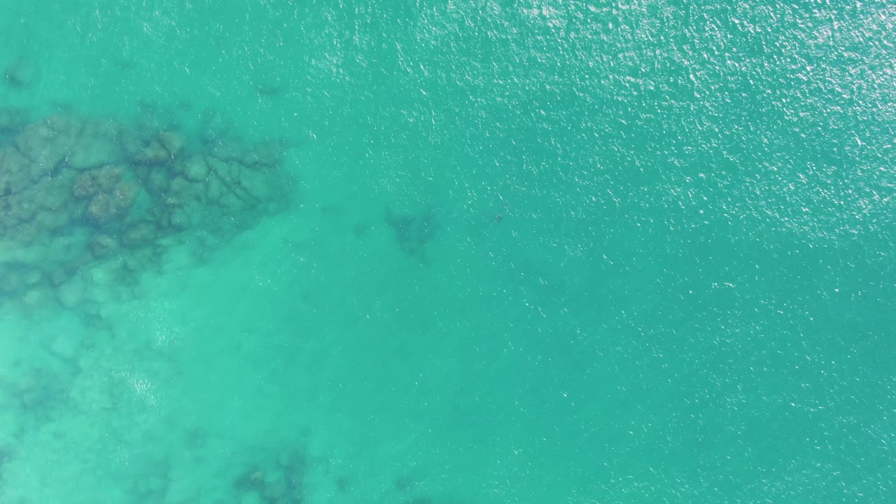Top-down Buzzards Beach frame with open turquoise water, faint ripples, and submerged rocks set back toward the upper left, with no visible people.