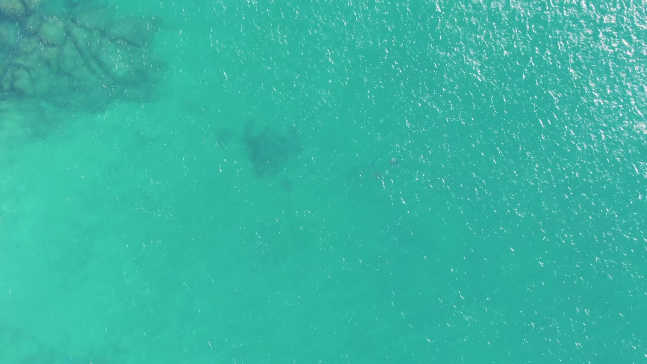 Top-down aerial frame of shallow turquoise water at Buzzards Beach, Baja California Sur, with faint darker underwater patches, bright ripples, and no visible people.
