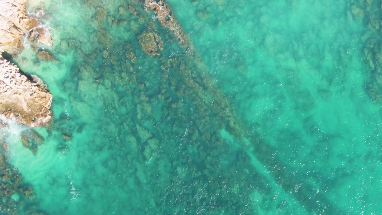 Overhead view at Buzzards Beach, Baja California Sur, showing turquoise water, submerged rock patches, pale sand at the left edge, and no visible people.