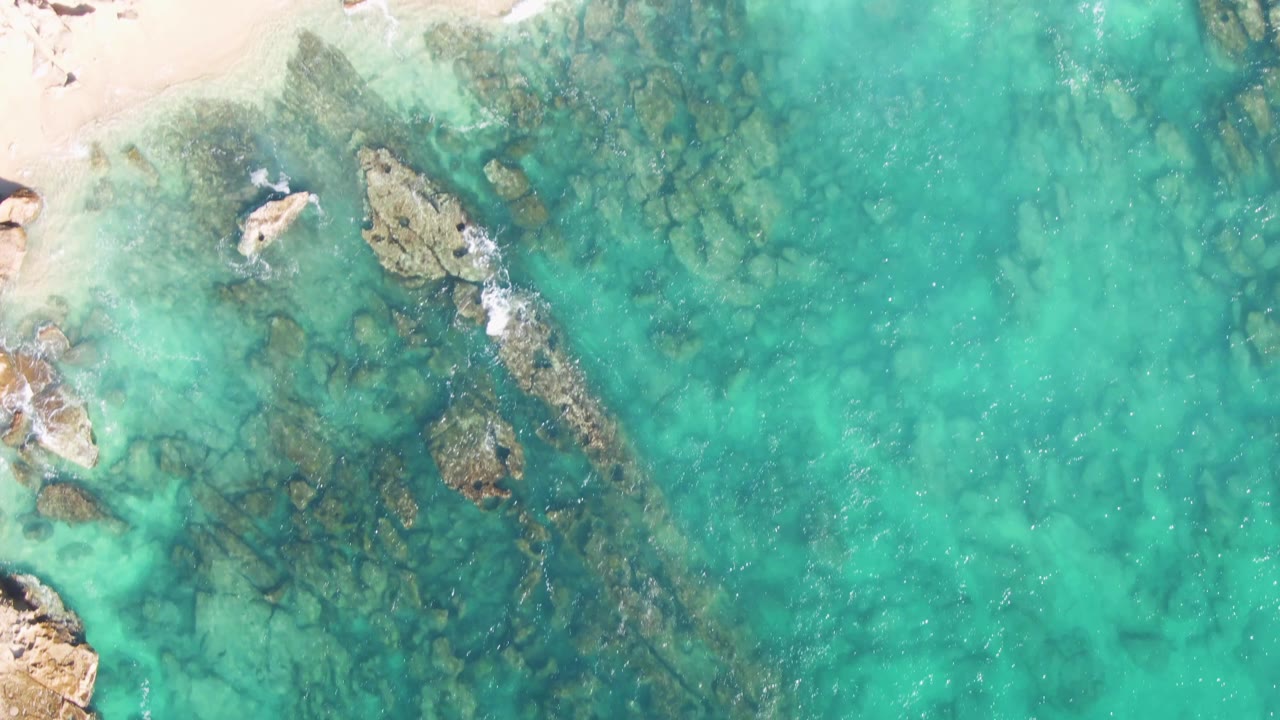 Overhead view of Buzzards Beach with pale sand across the top, rocks near the shore, clear turquoise water, and no visible people in this frame.