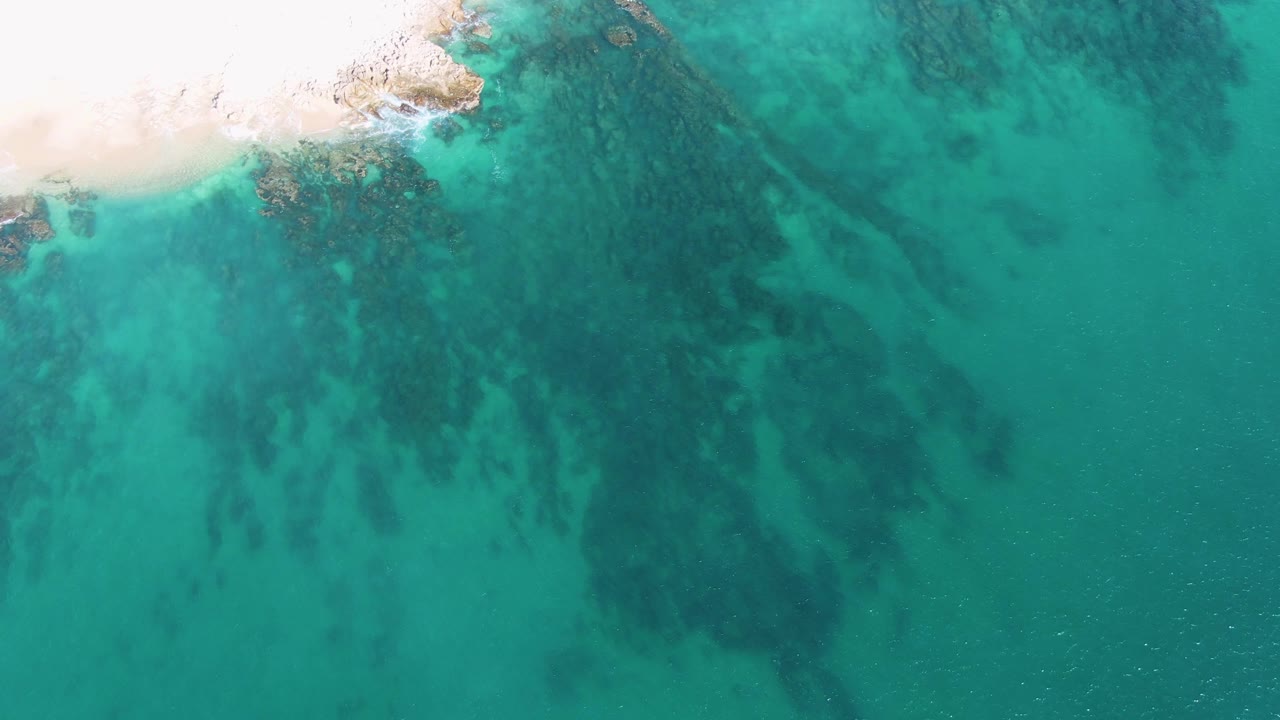 Overhead view of Buzzards Beach turquoise shallows with pale sand entering from the upper left, dark submerged rocks below the surface, and no visible people.