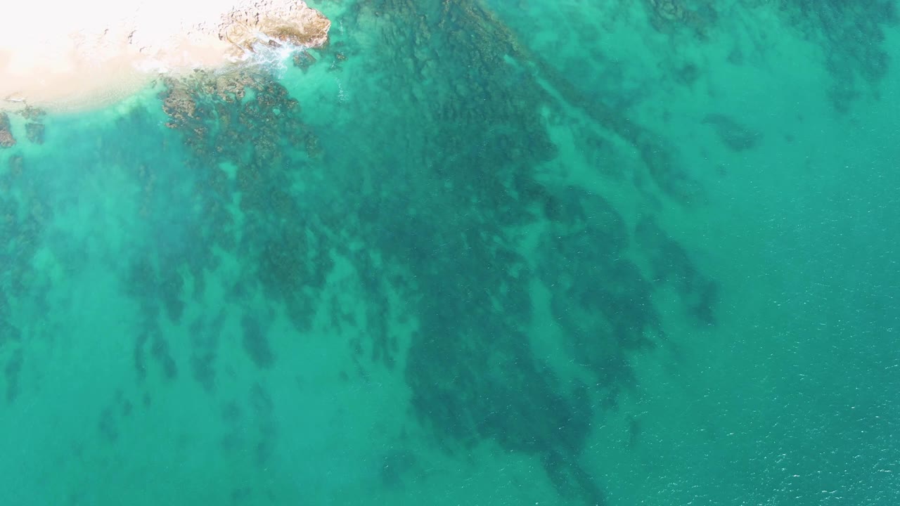 Aerial frame over Buzzards Beach showing a thin sandy shoreline at the top left, turquoise water across the frame, submerged rocky outcrops, and no visible people.