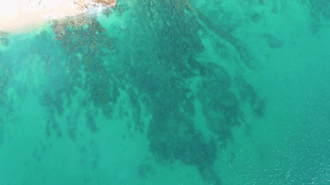 Overhead Buzzards Beach frame with a bright sand corner at the upper left, large darker submerged rock formations through the turquoise water, and no visible people.