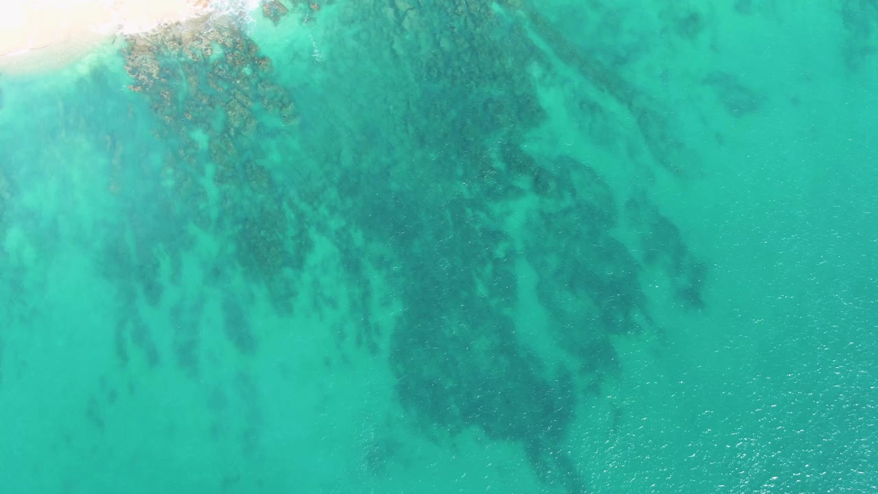 Overhead view at Buzzards Beach with only a small sand strip near the upper left, extensive turquoise shallows, mottled underwater rocks, and no visible people.
